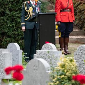 La gouverneure générale Julie Payette se tient à un podium.  Au premier plan, des pierres tombales blanches.  Derrière elle, à sa droite, un agent de police montée en rouge se tient au garde-à-vous.