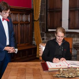 La gouverneure générale signe un livre d'or. Le maire de Mons, M. Elio di Rupo, est debout à sa droite et l'observe.