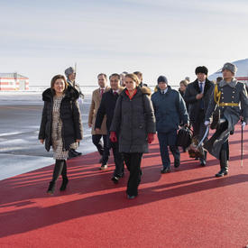 La gouverneure générale marche sur un tapis rouge qui mène à l'escalier d'un avion à l'arrière-plan.  Elle est entourée de représentants officiels.  Des gardes armés tapissent le tapis rouge.