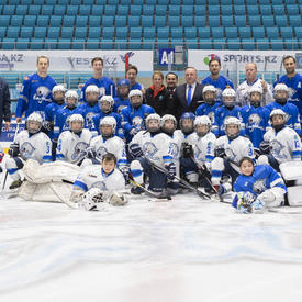 Deux équipes de hockey de jeunes posent sur la glace dans un aréna.  Les enfants portent des chandails blanc et bleu et de l'équipement de hockey. À l'arrière-plan, des adultes se tiennent debout, dont la gouverneure générale Julie Payette.