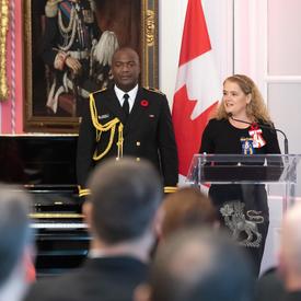 La gouverneure générale prononce une allocution debout devant un podium.  Derrière elle se trouve son aide de camp en uniforme de la Marine ainsi que le drapeau canadien.  