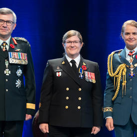 Le premier maître de 2e classe Marie Nathalie Isabelle Scalabrini se tient entre le Vice-chef d'état-major de la   Défense, le Lieutenant-général Paul Wynnyk (à gauche) et Son Excellence la très honorable Julie Payette (à   droite).