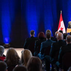  Le Vice-chef d'état-major de la Défense, le Lieutenant-général Paul Wynnyk, se tient devant un podium et s'adresse   à la foule, principalement en uniforme militaire Son Excellence la très honorable Julie Payette est assise et   écoute le discours.