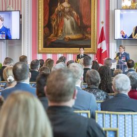 A photo of the room during the ceremony, with the Governor General speaking at the podium, displayed on two screens.