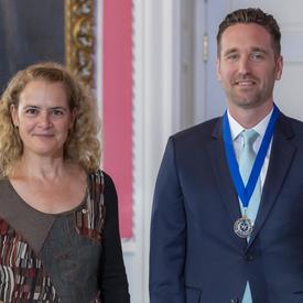 Governor General of Canada Julie Payette and reporter Corbett Hancey, wearing a medal with a blue ribbon, are standing looking directly at the camera.