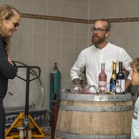 La gouverneure générale discute avec une jeune fille et Laurence-Olivier Brassard à l'intérieur du vignobles, où des bouteilles de vin sont montées sur un tonneau.