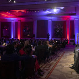 Vue de côté d'une salle où la gouverneure générale Julie Payette est vue, à l'extrême droite de la photo, sur scène, à un podium. La pièce est très sombre mais il y a des lumières violettes et roses.