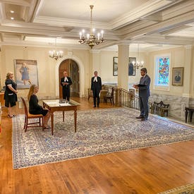 Side view of the Governor General sitting at a table. Behind her is a Canadian flag. In front of her, a man in a suit and tie is holding a document.