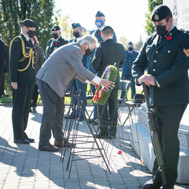 Mr. Whit Grant Fraser is laying a wreath in front of a stone memorial. Several members of the Canadian Armed Forces are standing around him, wearing masks. Behind him, there is a man in a black coat standing at a podium. They are outside and it is sunny.