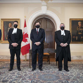L’administrateur se tient entre quatre personnes. Toutes les personnes portent des masques. Un drapeax du Canada se trouve à l’arrière-plan.