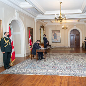 Quatre personnes se tiennent debout à une certaine distance pendant que l’administrateur, assis à une table, signe un document.