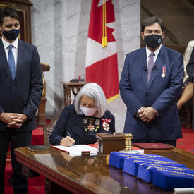 Mary May Simon signing a document in the Senate. The Prime Minister and the Supreme Court Chief Justice Richard Wagner stand on either side of her. 
