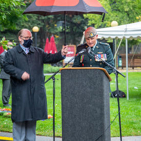 A man in uniform speaks at a podium. Another man holds an umbrella to protect him from the rain.