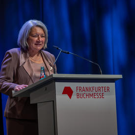 La gouverneure générale Mary May Simon se tient derrière un podium gris. Elle porte un tailleur rose. Derrière elle se trouve un rideau bleu foncé. Sur le podium, en rouge, on peut lire «Frankfurter Buchmesse».