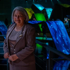La gouverneure générale Mary May Simon se tient devant une grande exposition artistique illuminée. Celle-ci est composée de rampes bleues, vertes et jaunes. Elle sourit. 