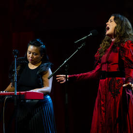 Iskwē and an accompanying musician are on a stage. Iskwē is wearing a red dress and singing into a microphone. Her accompanying musician is wearing a black dress and is sitting behind a keyboard playing a song.