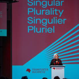 The Governor General is standing behind a grey podium. She is speaking into a microphone. On the podium, there is red text that reads, “Frankfurter Buchmesse.” Behind her a screen reads "Singular Plurality, Singulier Pluriel."