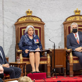 Their Excellencies seated at a pair of thrones. Her Excellency holds the binder containing the Throne speech. The Prime Minister is off to her right.