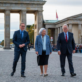 Mr. Michael Müller, the Governing Mayor of Berlin, Governor General Mary Simon and Mr. Whit Grant Fraser. They are smiling and standing on the street in front of the Brandenburg Gate. There is a crowd gathered along the street behind them.