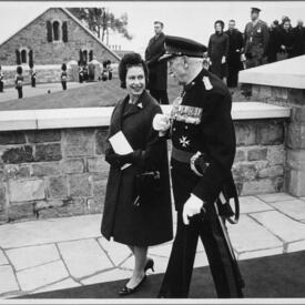 Une photo en noir et blanc de la Reine qui marche avec le gouverneur général Vanier. Ils se promènent le long des remparts de pierre de la Citadelle. On voit à l’arrière-plan une petite foule, des gardes en uniforme et un bâtiment en pierre.