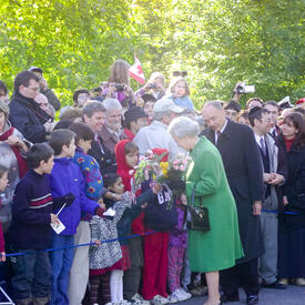 La Reine, qui tient des bouquets de fleurs et porte un manteau vert, salue une foule à l’extérieur de Rideau Hall.