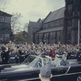 La Reine et le duc d’Édimbourg sont conduits dans une décapotable noire. Une grande foule les salue. On voit une cathédrale à l’arrière-plan.