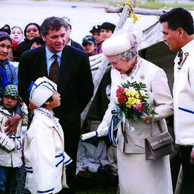 La Reine, vêtue de blanc avec un bouquet de fleurs à la main, est accueillie par un enfant qui porte un manteau et un chapeau de cérémonie blanc et bleu. Deux membres du personnel regardent la scène. On voit une foule derrière eux.