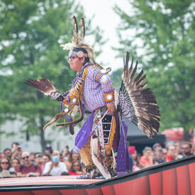Un artiste autochtone danse sur la scène, portant un costume mauve avec des plumes lors de la cérémonie de la fête du Canada.