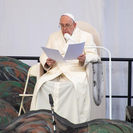 Le pape François prononce un discours. Il est assis sur une chaise blanche et tient des feuilles de papier dans ses mains.