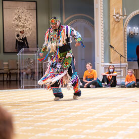 A dancer performing in the Ballroom at Rideau Hall. He is wearing a traditional outfit. Schoolchildren are seated around him, watching.