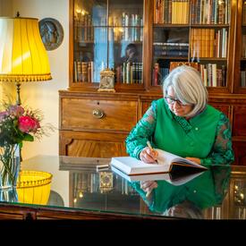 Governor General Mary Simon is sitting at a desk in a study. She is writing in a book. 