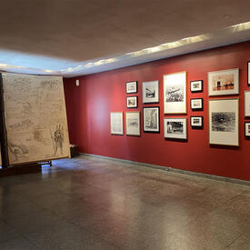 Il y a une grande exposition de 2 pièces de croquis et de textes sur la gauche. Sur la droite, un mur rouge présente un mélange de 17 photographies, croquis et peintures.