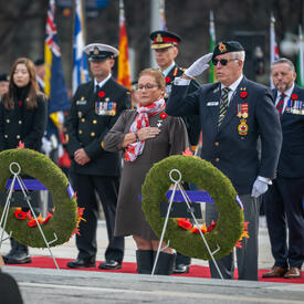 Mrs. Candy Greff is standing in front of two wreaths at the National War Memorial in Ottawa. A man is standing beside her, saluting. Several people are standing behind them.