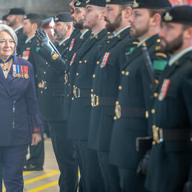 Governor General Mary Simon, wearing a purple suit and military medals, walks in front of a guard of honour composed of members wearing dark green uniforms.
