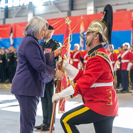 Vue de profil de la gouverneure générale Mary Simon, portant un complet violet, qui donne un drapeau sur un mât à un militaire en uniforme rouge agenouillé devant elle.