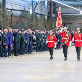 Trois militaires en uniforme rouge, portant des gants blancs et des casques dorés, marchent devant 3 lignées de spectateurs debout. Le militaire du centre porte un drapeau rouge sur un mât.