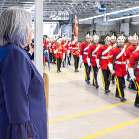 Governor General Mary Simon, wearing a purple jacket, stands and watches as a guard of honour walks in front of her.