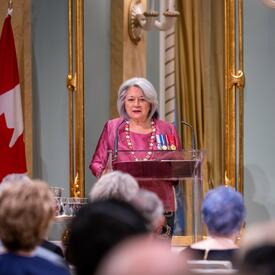 La gouverneure générale Mary Simon porte un habit rose. Elle se tient derrière un pupitre devant une grande foule dans la salle de bal de Rideau Hall. Derrière elle, on voit un grand miroir et un drapeau du Canada.