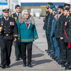 La gouverneure générale Simon marche à l'extérieur. Un homme en uniforme militaire marche à ses côtés, à sa gauche. À sa droite, il y a une rangée d'individus en uniforme militaire.