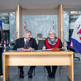 La gouverneure générale Simon et M. Fraser assis à une table en bois. L'honorable Caroline Cochrane se tient à leur droite et sourit à la caméra.