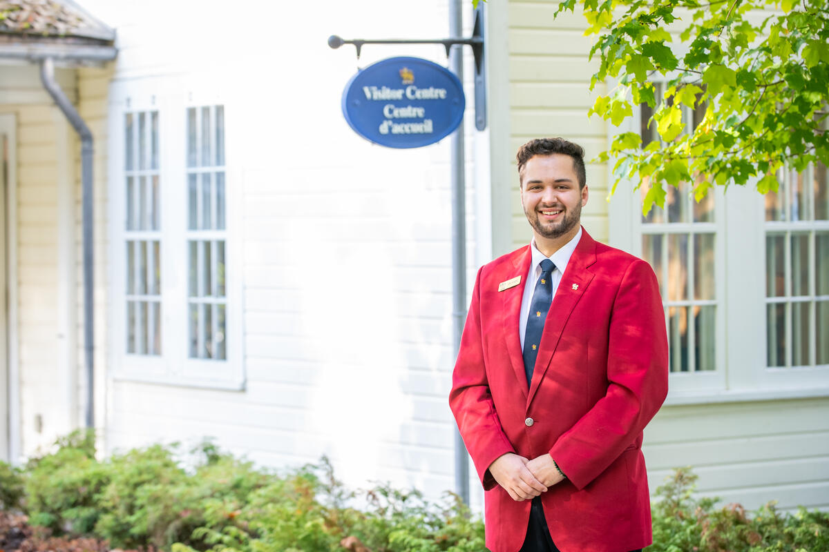 Un guide vêtu d'une veste rouge se tient devant un bâtiment sur lequel est accrochée une pancarte indiquant « Visitor Centre / Centre d'accueil ».