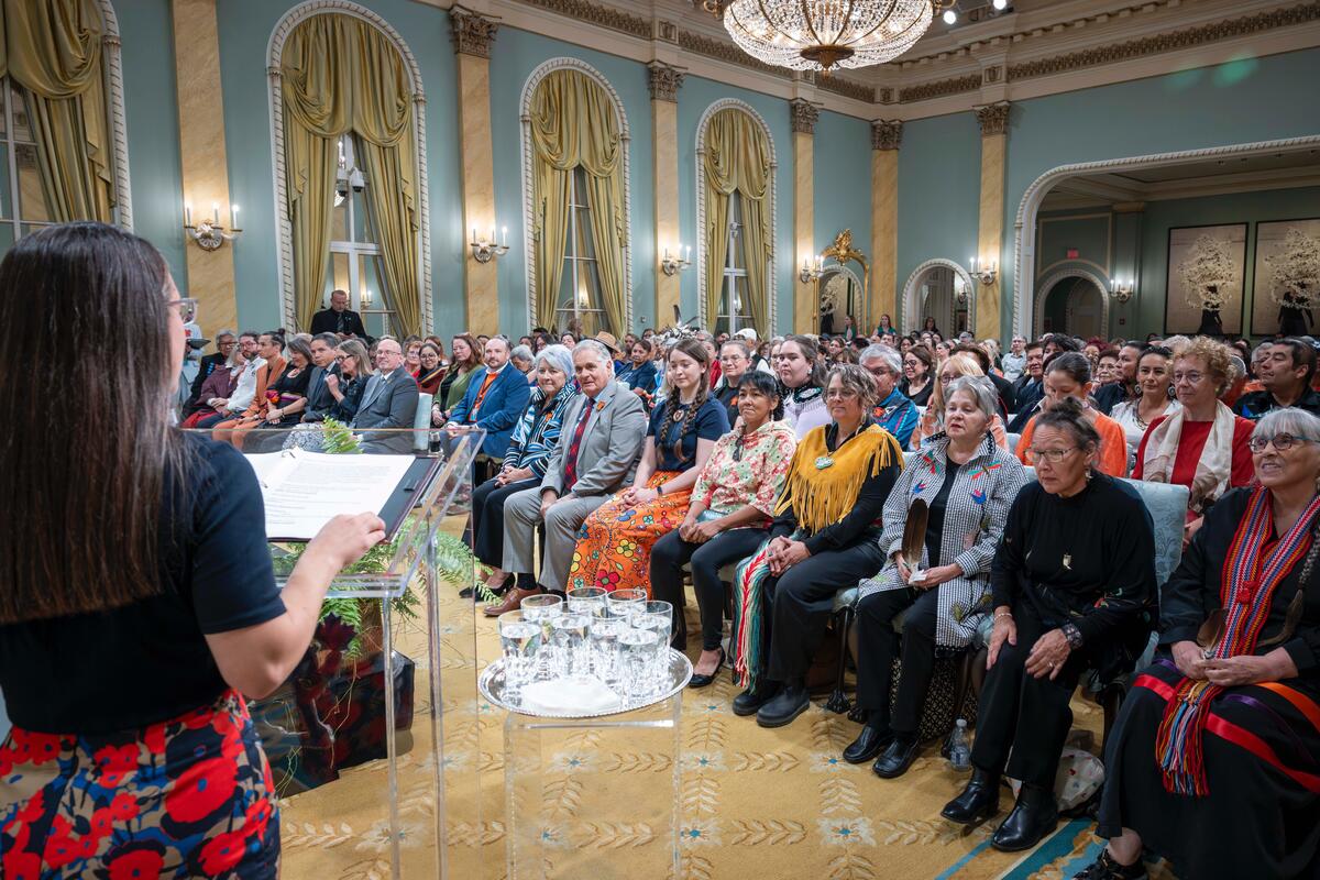 A speaker addresses a group of listeners from a podium. Some are wearing Indigenous regalia.