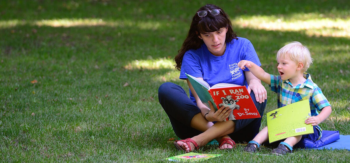 Une femme lit un livre à un petit garçon. Ils sont assis sur une couverture bleue sur l'herbe à l'extérieur.