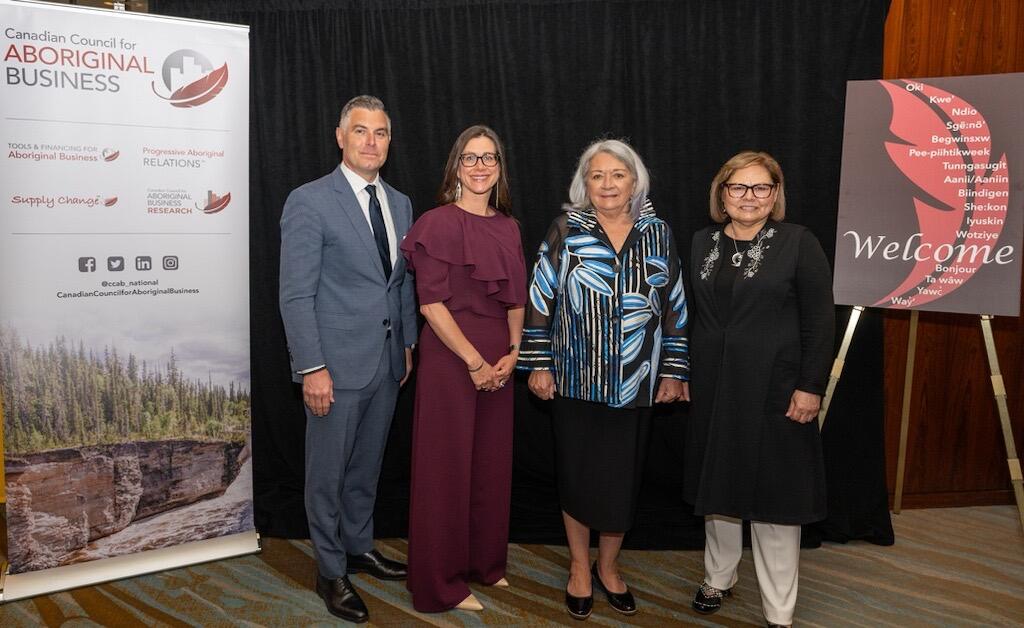 Governor General Mary Simon posing for a picture with three people. There is a banner on the left and an easel with a welcome sign on the right. 