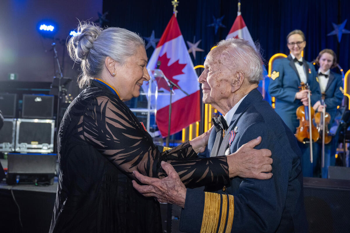 La gouverneure générale Mary Simon tient le général Richard Rohmer dans ses bras pendant que les membres de la Musique centrale des Forces canadiennes regardent en arrière-plan