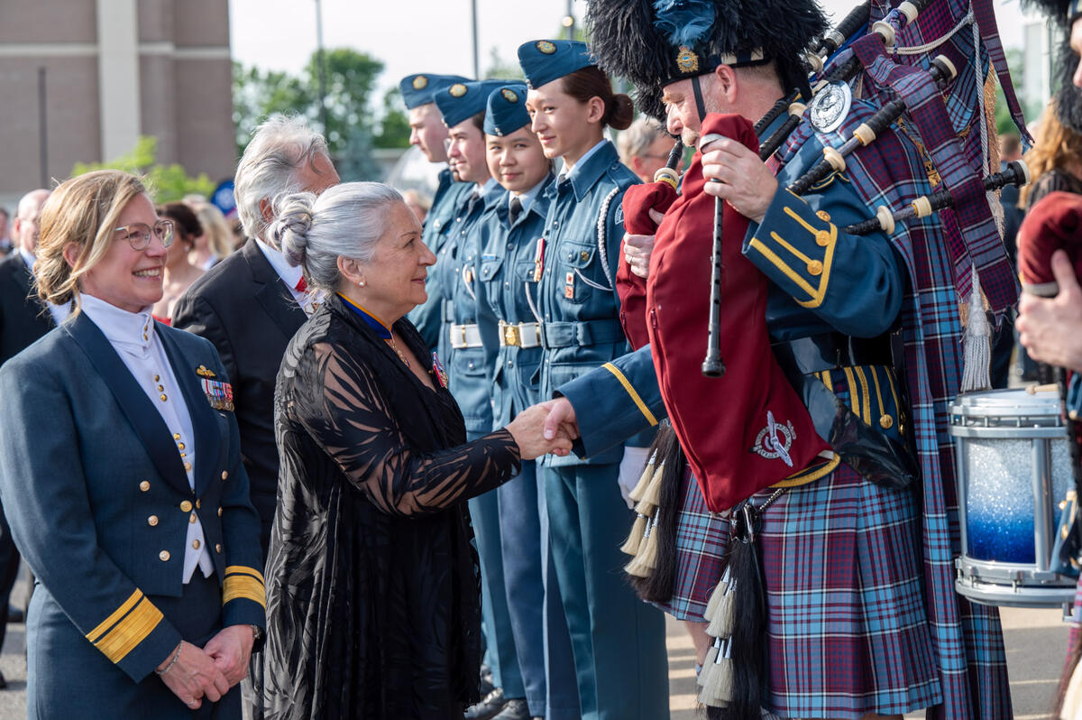Governor General Mary Simon is shaking hands with a piper. Several people wearing blue military uniforms appear in the background.