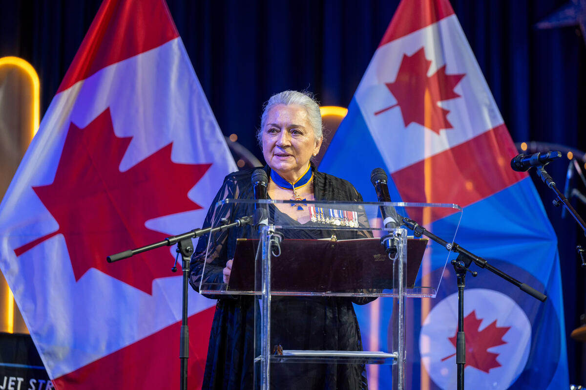 Governor General Mary Simon is standing behind a clear podium. She is speaking. The Canadian flag and a flag representing the Royal Canadian Air Force are displayed behind her.