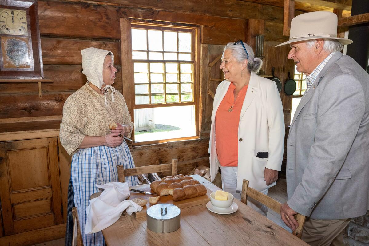 La gouverneure générale Simon et M. Whit Fraser s'entretiennent avec une personne costumée dans une vieille ferme. Ils sont debout autour d'une table.