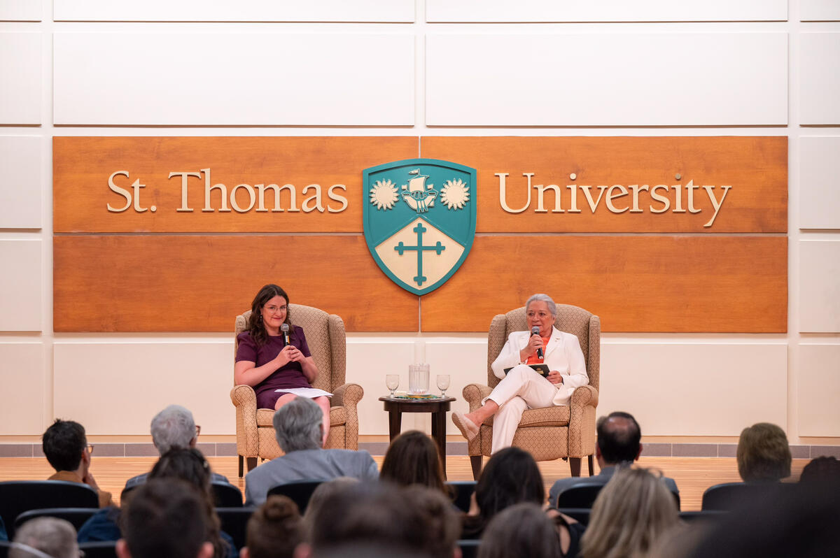 Governor General Simon and another person sitting in lounge chairs on stage. Both have a microphone. The words “St. Thomas University” are on the wall behind them.