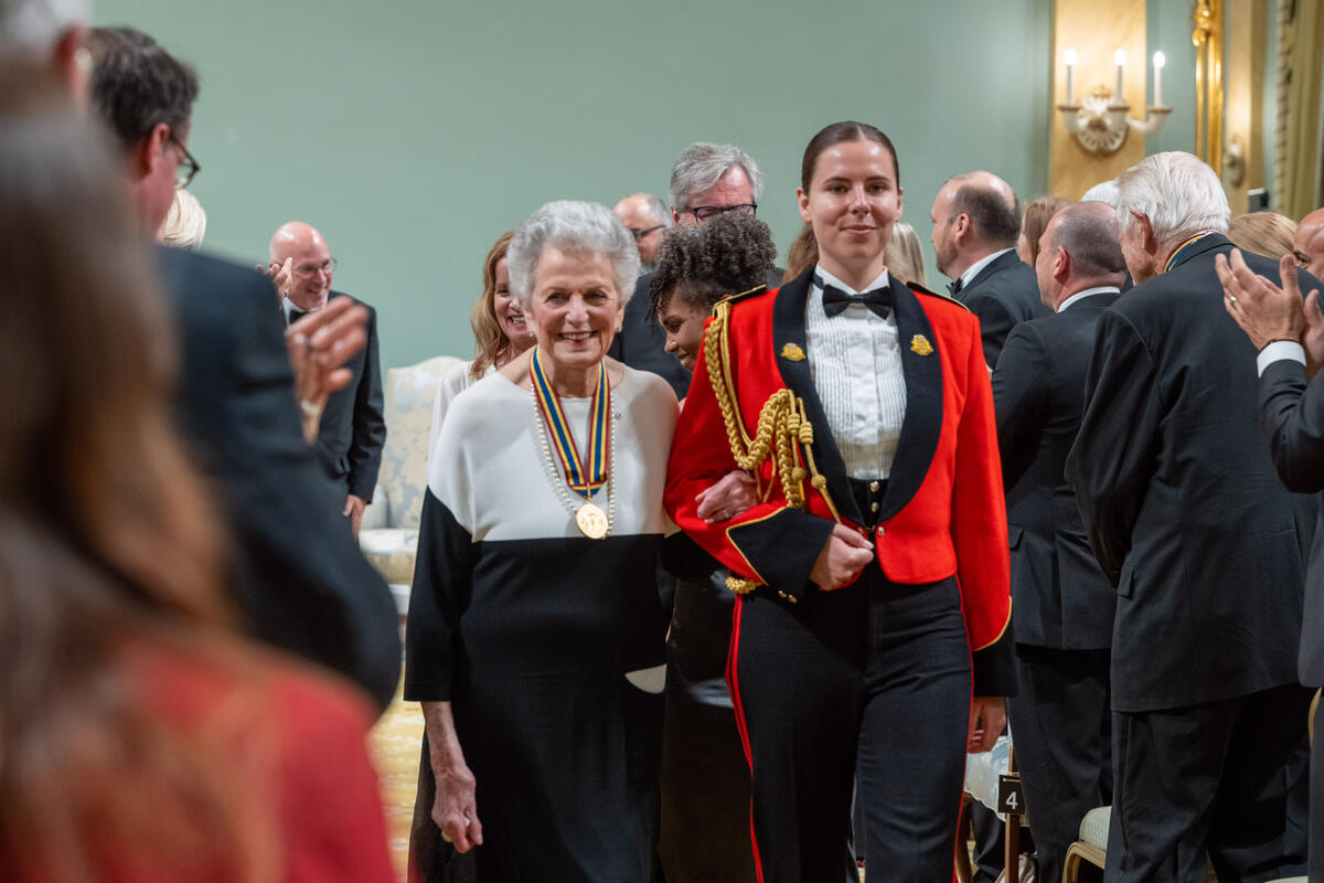Jenny Belzberg, accompagnée d'un membre des Forces armées canadiennes en uniforme rouge, sort de la salle de bal de Rideau Hall à travers le public